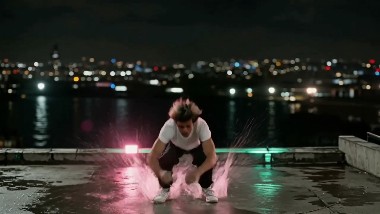 A dancer on a wet rooftop drives into a tight tucked backflip and lands on both feet in a deep squat with a splash
