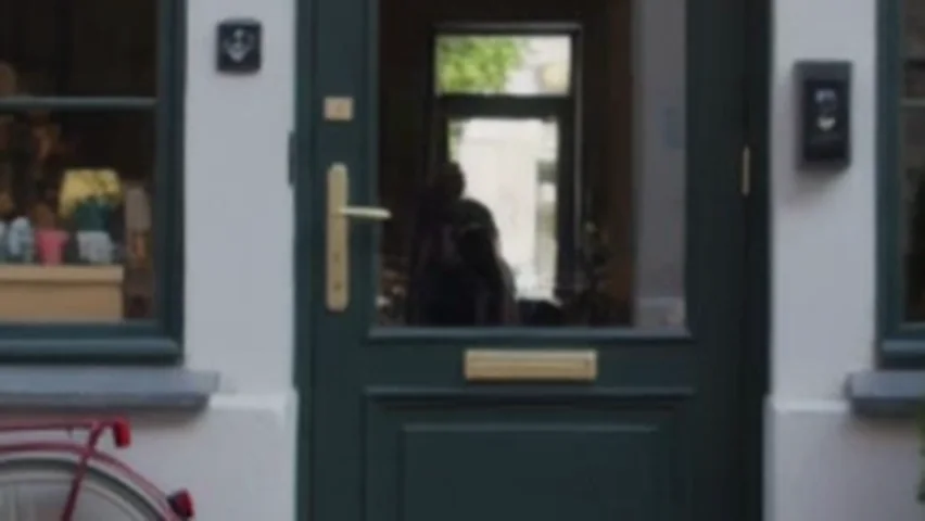 A close crop of Kling v3's first run, enlarged: a dark figure stands inside the bakery, seen through the glass of the green door.