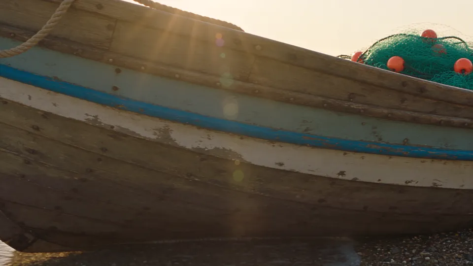 Seedance 2.5 at 1080p: an old blue and white fishing boat on a pebble beach, backlit by a low sun